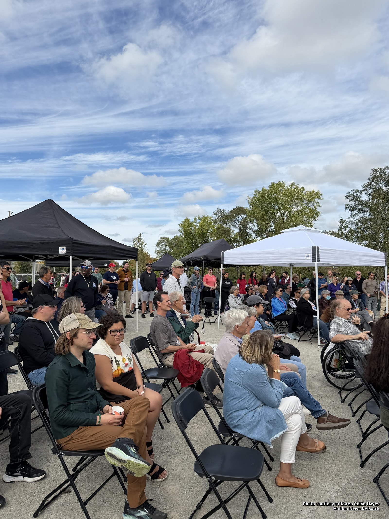 A crowd seated on folding chairs and standing under black and white pop-up tents at the Malden River Works grand opening, under a blue sky with scattered clouds.
