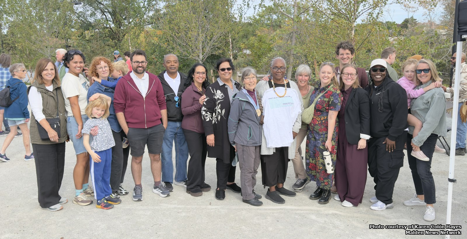 About twenty adults and several children standing in a line for a group photo on a gravel plaza, with trees behind them.