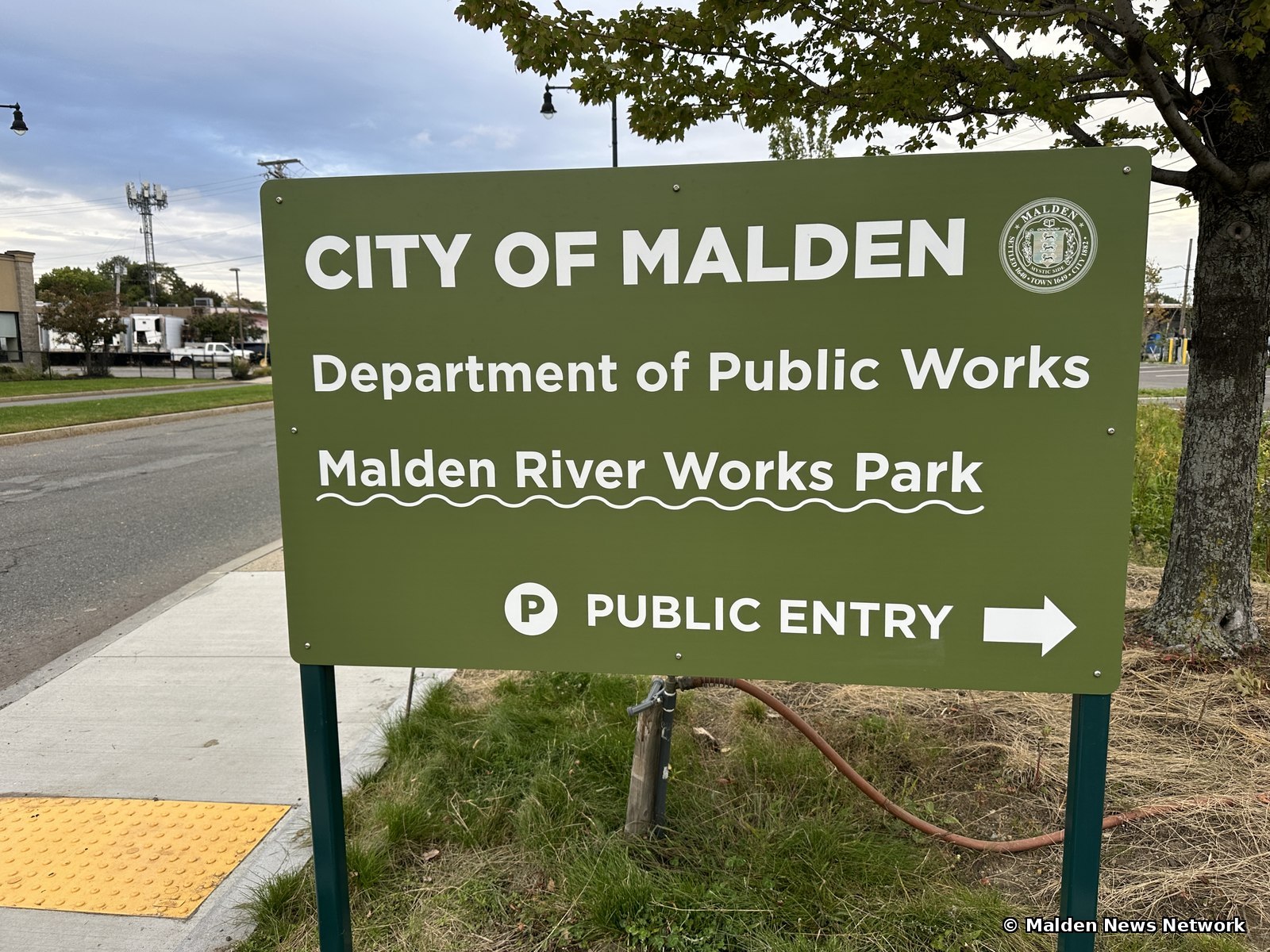 A green City of Malden Department of Public Works sign reading Malden River Works Park, Public Entry, with an arrow pointing right.
