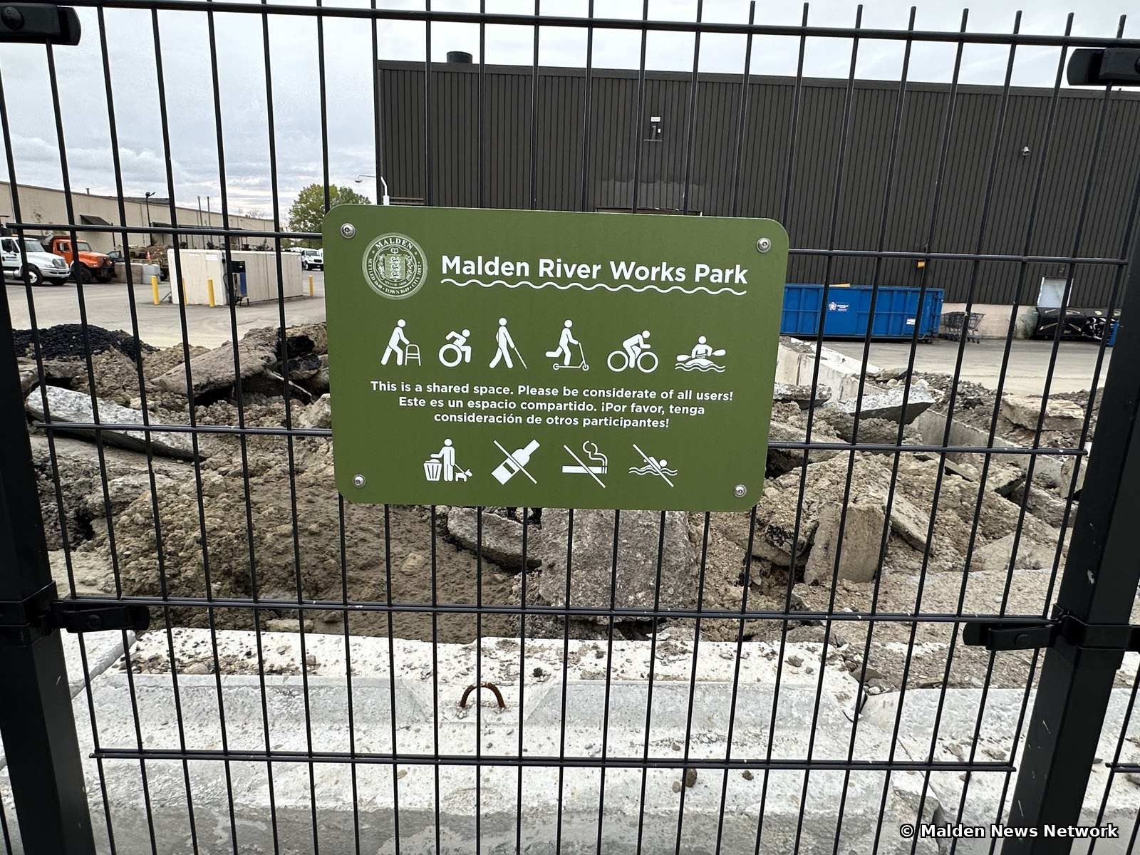 A green park sign on a black fence asking visitors to be considerate of all users, with a DPW yard and piles of broken concrete and soil visible behind it.