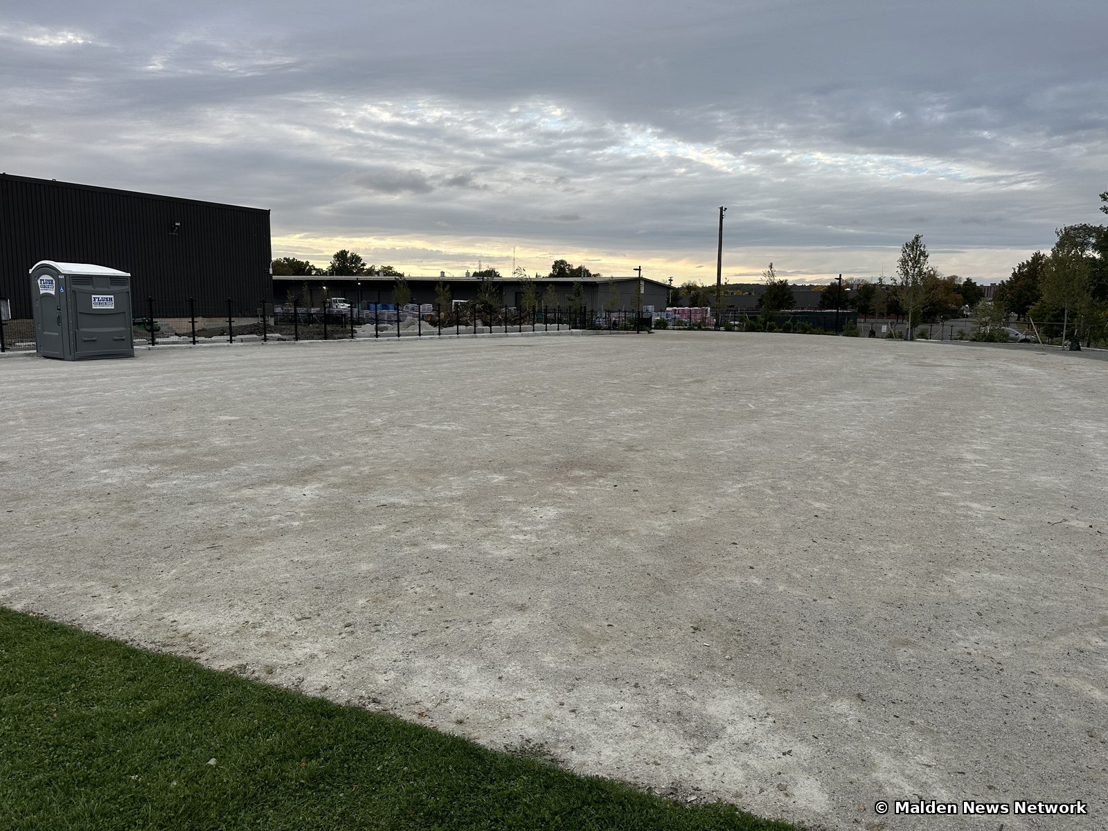 A small strip of lawn in the foreground beside a very large sandy gravel expanse with a portable toilet and dark industrial buildings beyond.