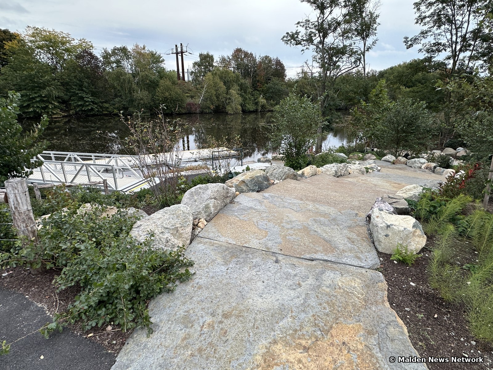 A walkway of large flat stone slabs bordered by boulders leading toward a floating dock on the river, with no handrail in view.