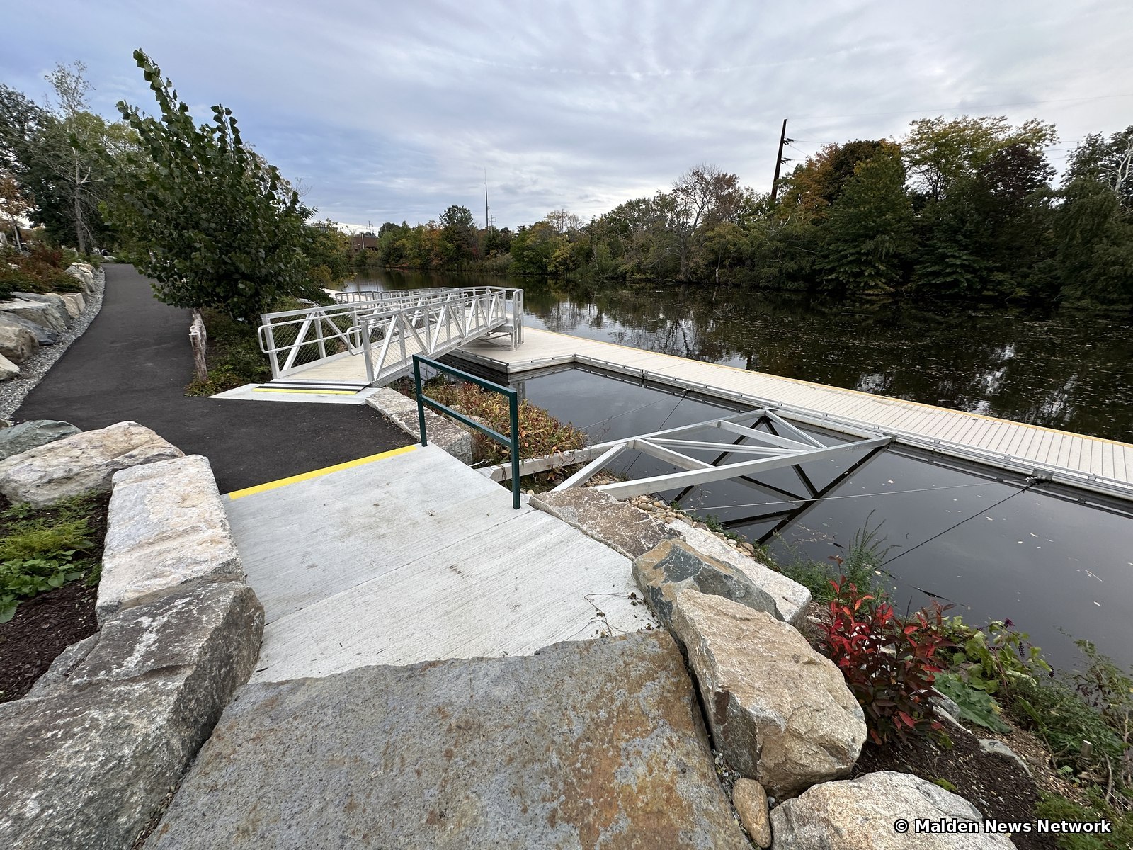 A concrete landing, aluminum gangway, and long floating dock on the Malden River, with boulders and red-leaved plants in the foreground.
