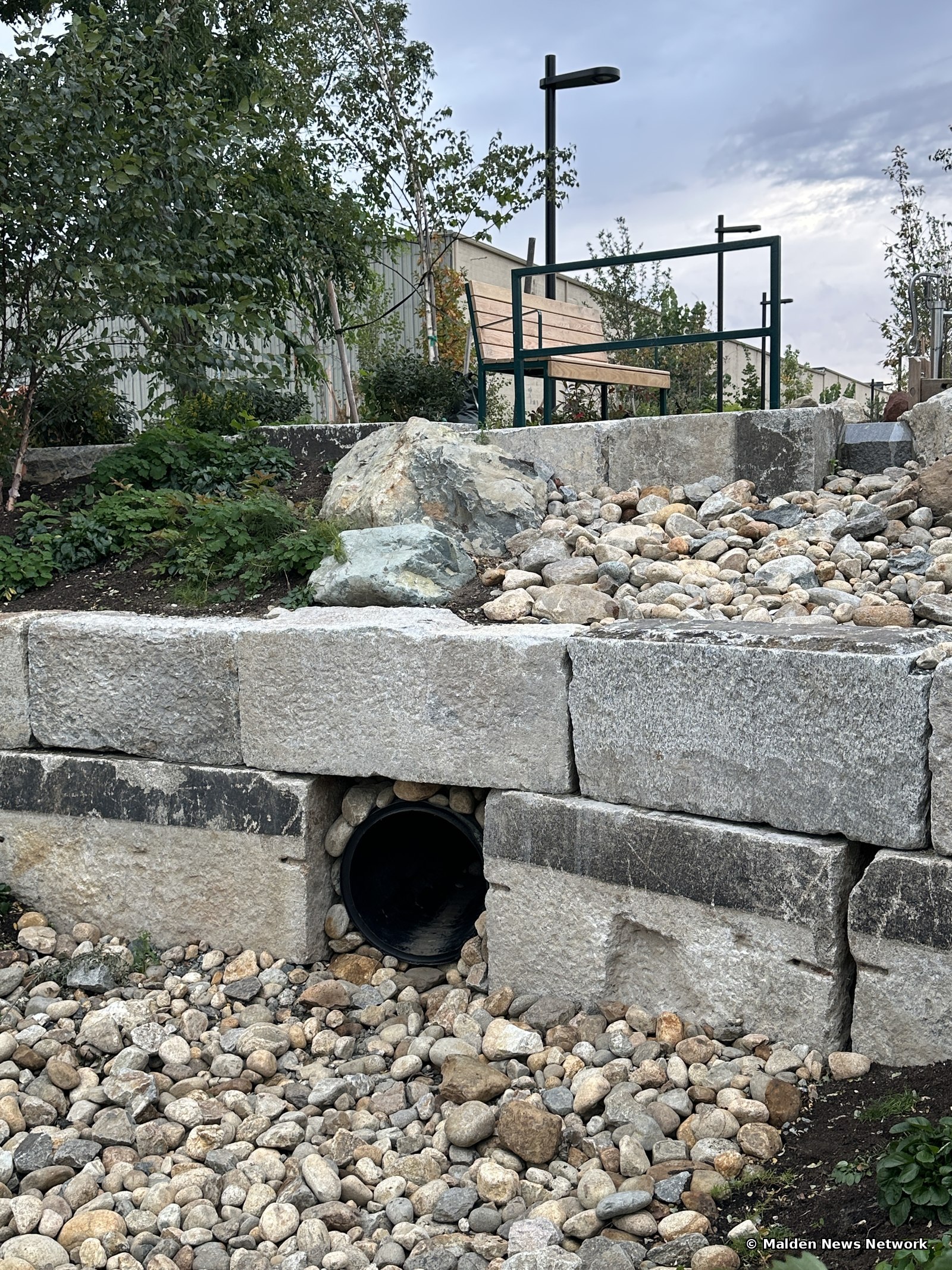 A black culvert pipe opening set into a stone block retaining wall above a bed of river stones, with a bench and light pole on the terrace above.
