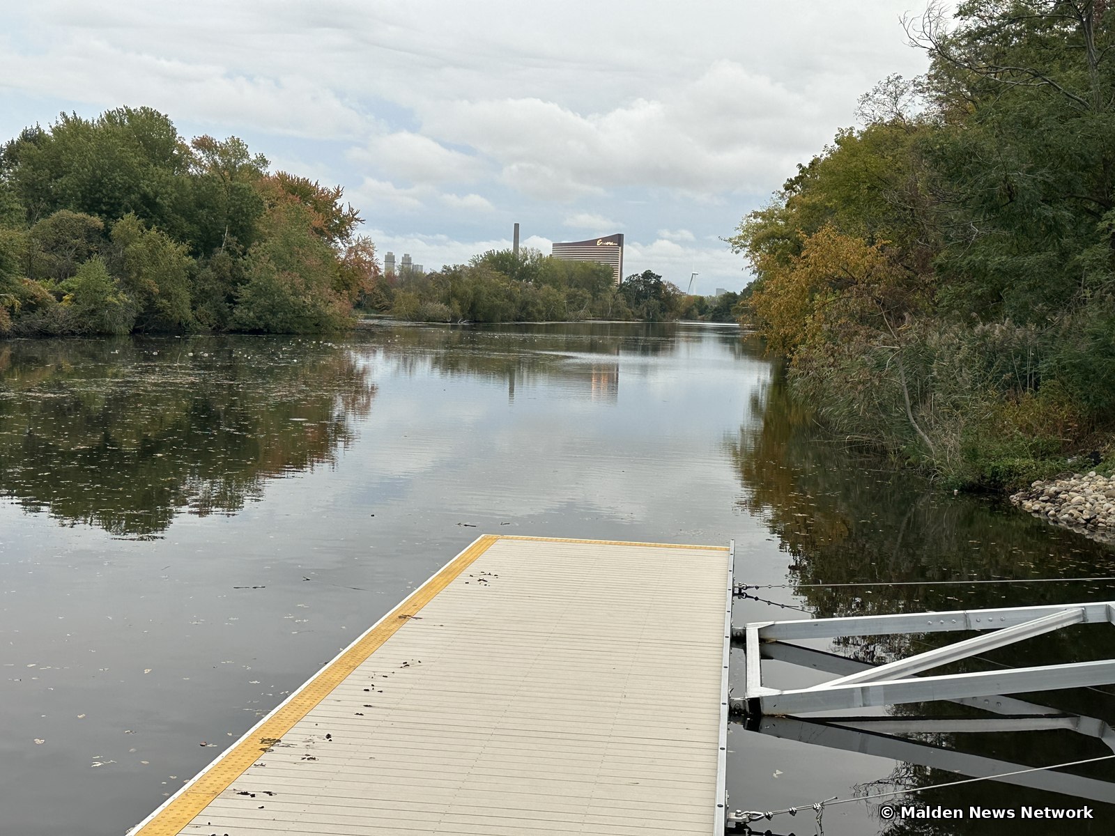 The end of a floating dock on a calm stretch of the Malden River, with autumn-colored trees on both banks and a distant tall building on the horizon.
