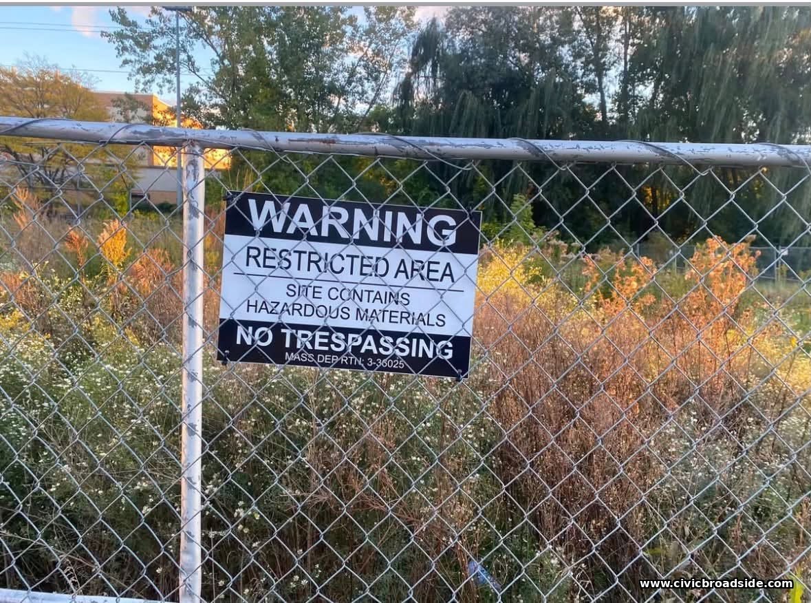 Warning sign on the perimeter fence reading 'Restricted Area, Site Contains Hazardous Materials, No Trespassing'