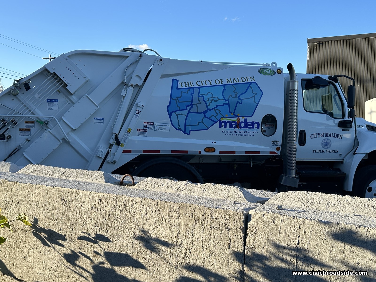 A white City of Malden Public Works rear-loader truck with a map of the city on its door, seen over a rough stone wall