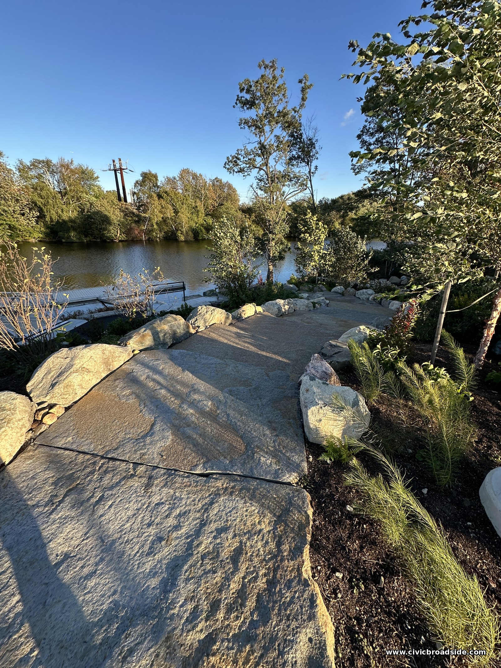 A flat stone path and boulders leading down toward the river, with young trees and grasses on both sides
