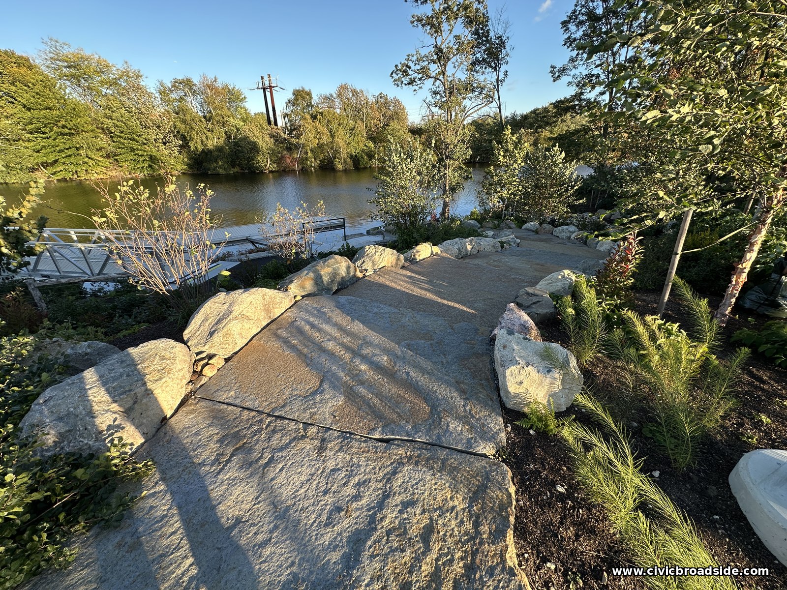 Broad irregular stone slabs descending between boulder edging and plantings, with long shadows and the river beyond