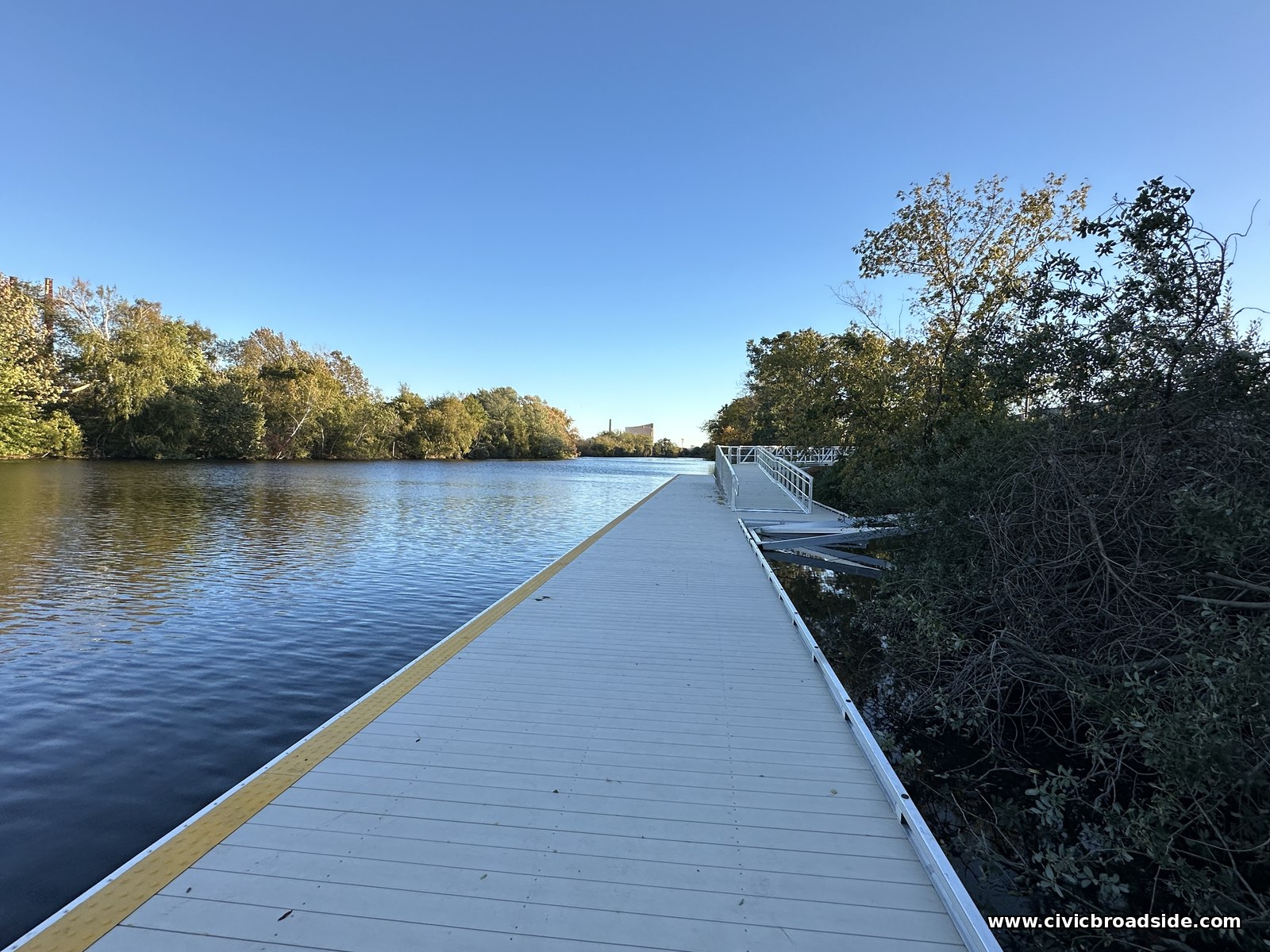 A long floating dock of pale decking with a yellow edge stripe running along open water, with a railed section at the far end