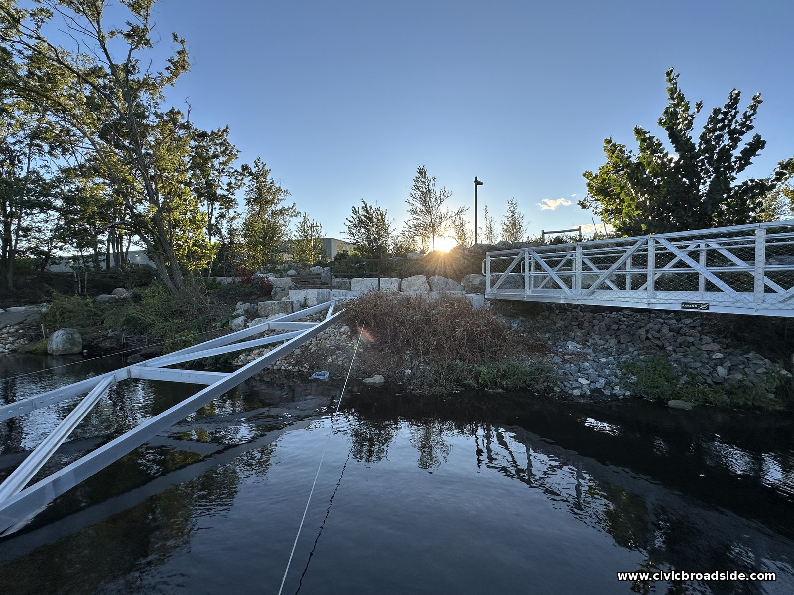 White aluminum struts angling down into dark water beside a white railed walkway, with the low sun and wind-bent trees behind