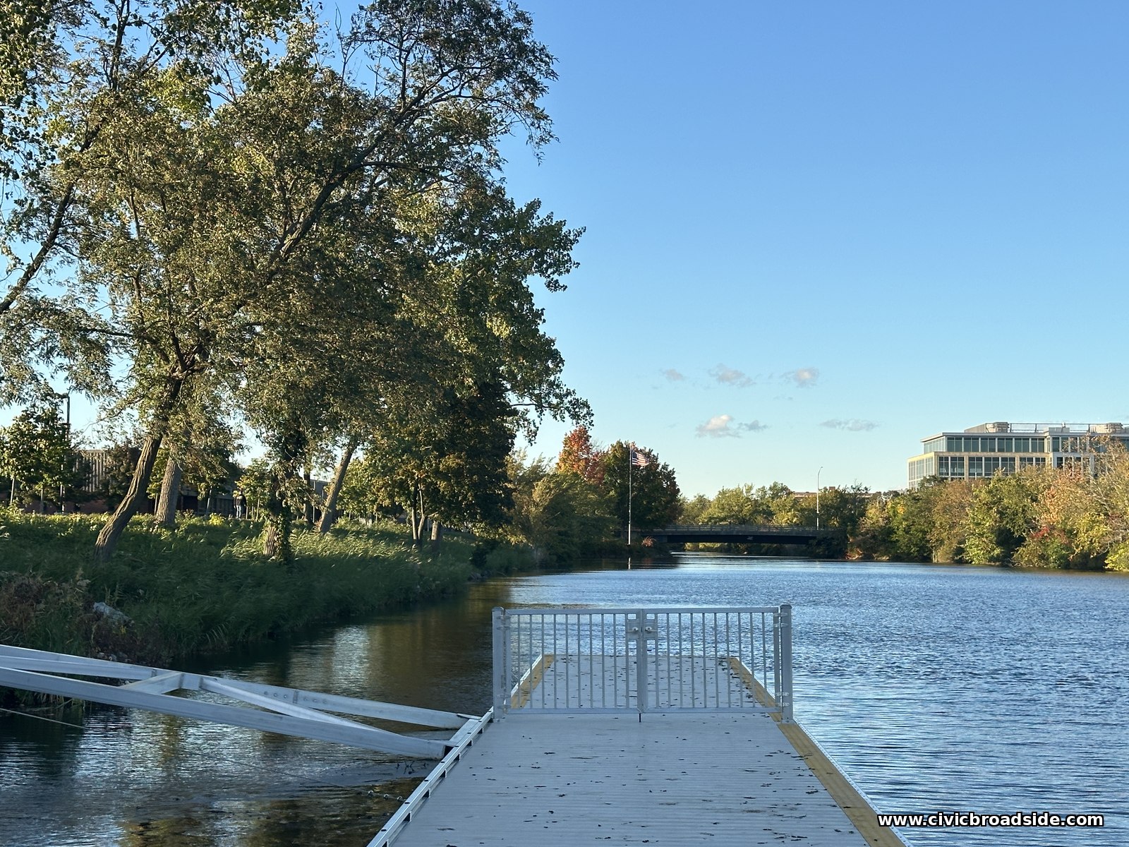 A small double gate of white aluminum bars across the far end of a floating dock, with open river ahead, an aluminum strut angling into the water at left, and a bridge and flag in the distance