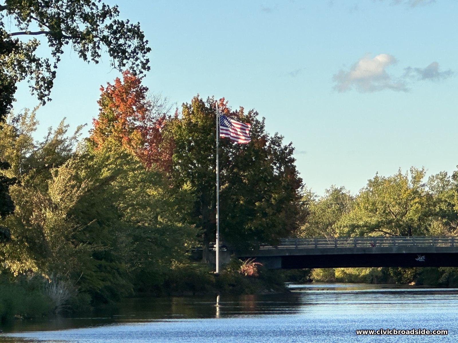 An American flag on a pole among trees turning red and gold, with a bridge crossing the river behind it