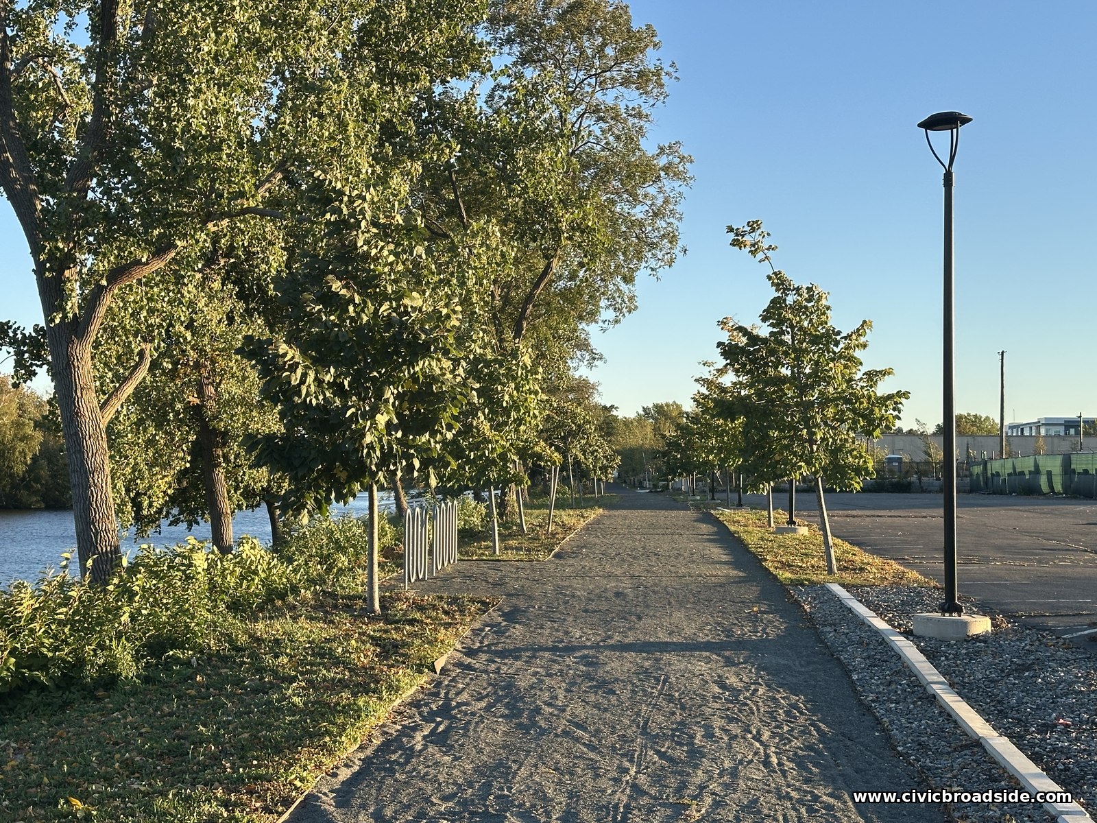 A stone-dust path beside the river lined with young trees and a tall black light pole