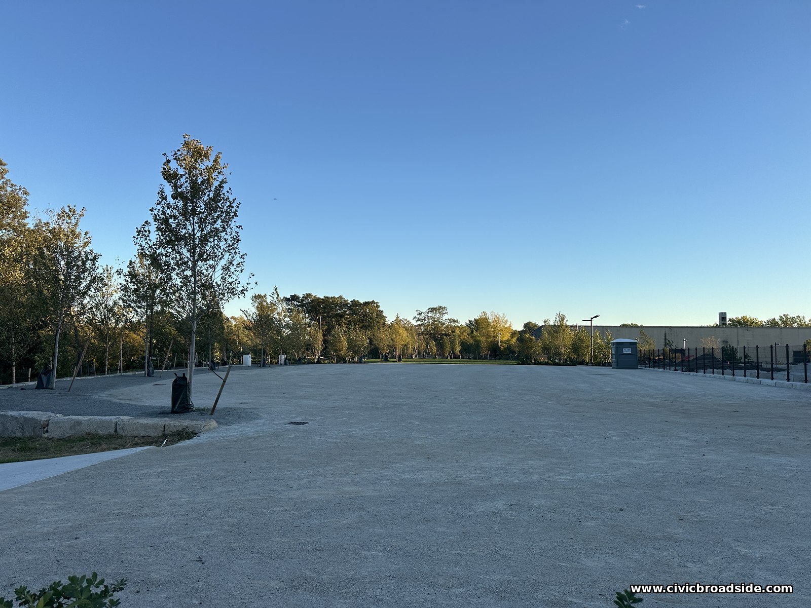 A broad open gravel area with a few young trees, a fence, and a portable toilet in the distance