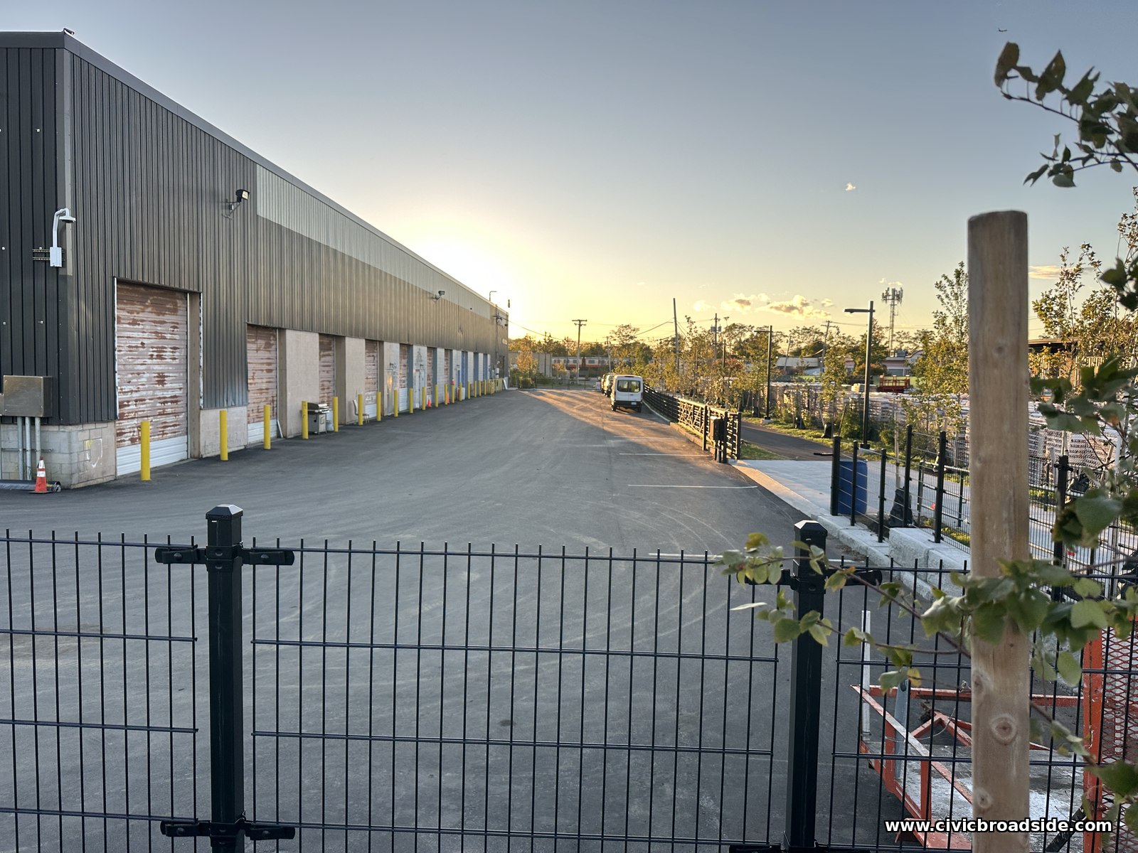 Late sun glowing at the end of a long fence line beside a grey building with loading bay doors
